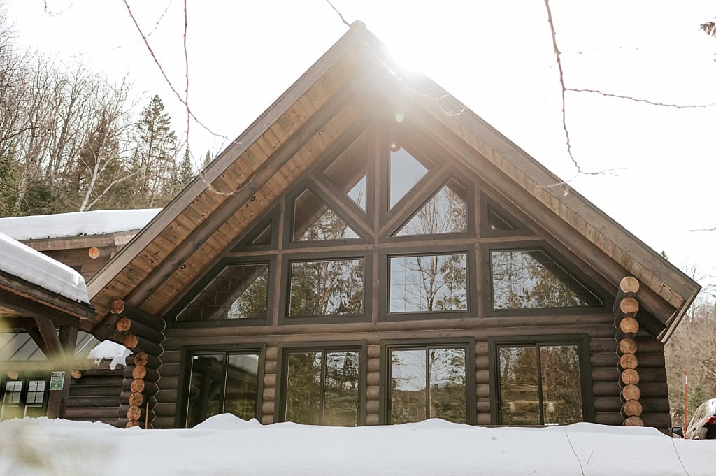 Exterior of a log cabin chalet in Mont-Tremblant with floor-to-ceiling triangular windows and a snow-covered roof, sunlight streaming through the trees in winter