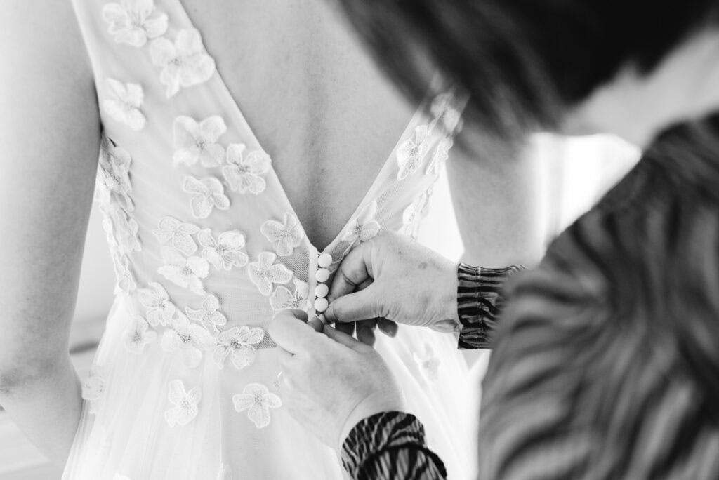 Black and white close-up of hands buttoning the floral appliqué back of a wedding dress on the morning of an intimate Mont-Tremblant wedding